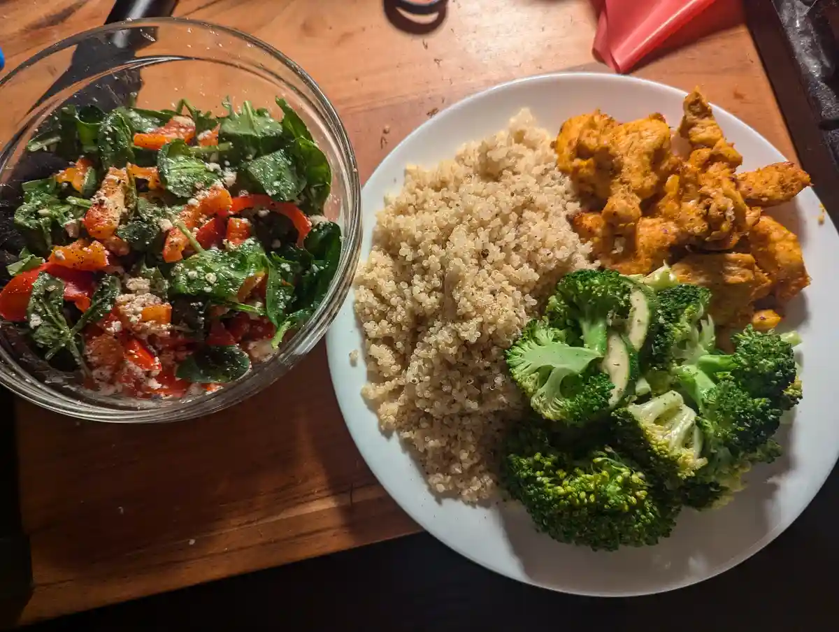 A top-down photo shows a white plate with cooked quinoa, seasoned chicken pieces, and steamed broccoli, next to a glass bowl…