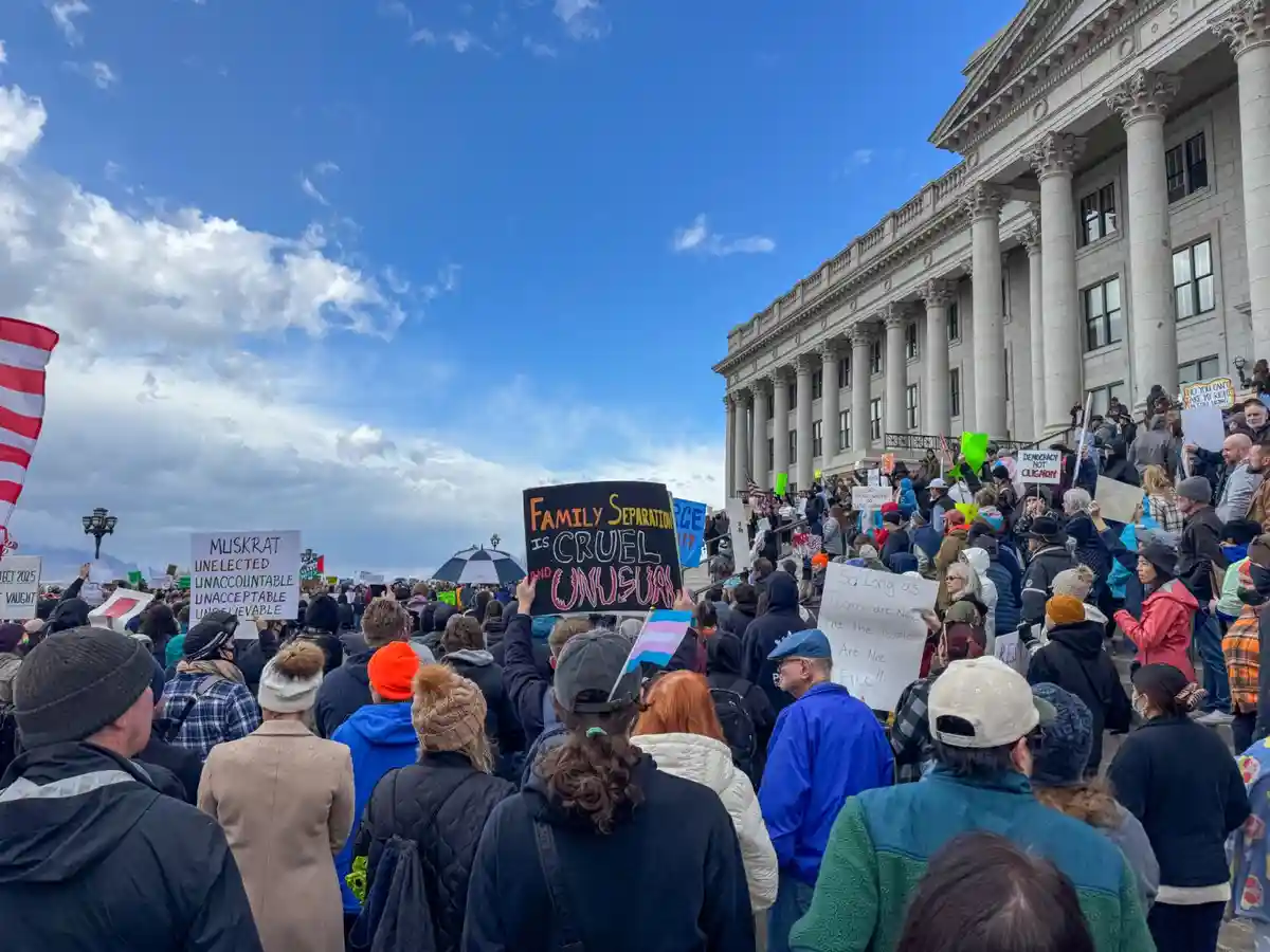 A large crowd of protesters gathers outside a grand neoclassical government building under a partly cloudy sky, holding…