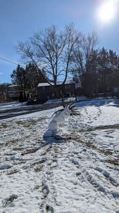 A small snowman with a smiling face and twig arms is leaning over in a snowy yard under a bright sun. The snowman appears to…