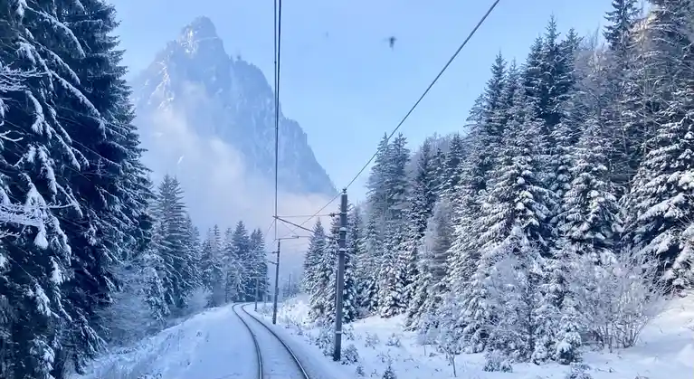 A snowy landscape viewed from a train, showing railway tracks curving through a forest of snow-covered evergreen trees with…