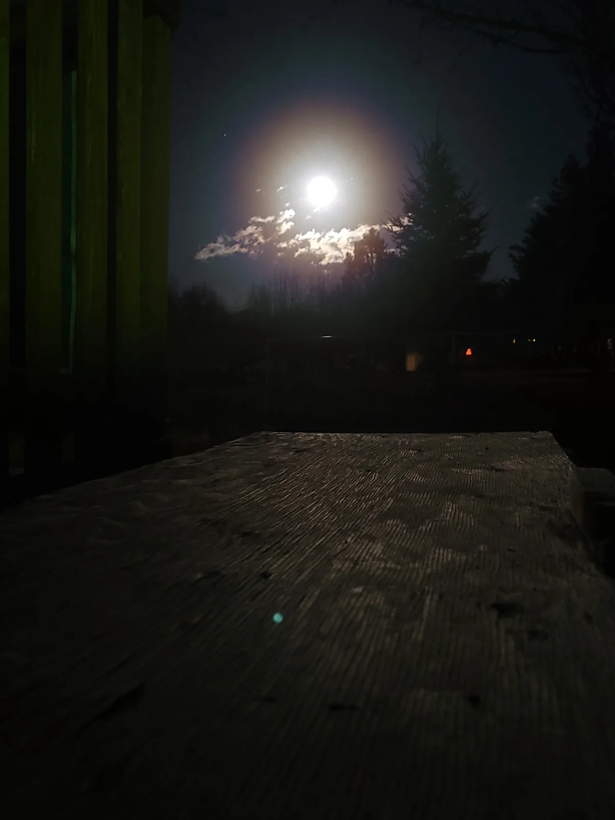 A low-angle nighttime photo shows a bright full moon partially obscured by clouds, viewed from a wooden deck with vertical…