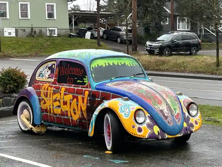 A vibrantly painted vintage Volkswagen Beetle is parked on a wet street, featuring psychedelic patterns, a brick wall design…