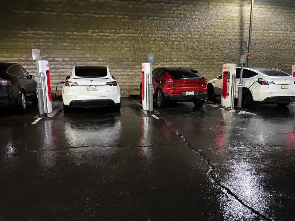 A row of electric vehicles, including a red Volvo and several Teslas, are parked at charging stations in a wet, dimly lit…
