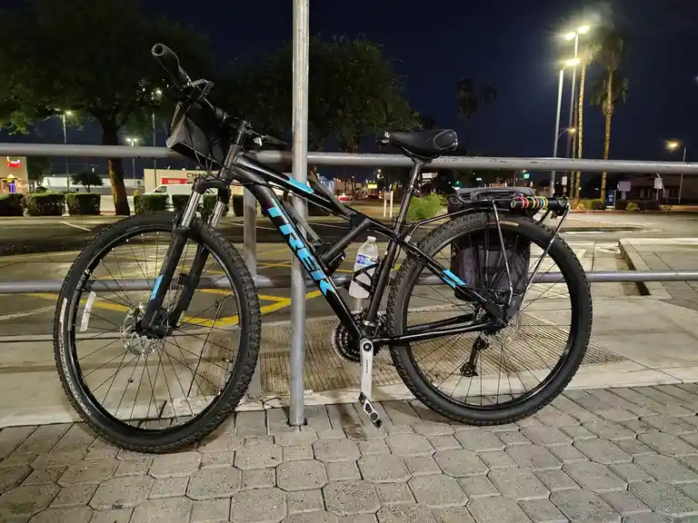 A black Trek mountain bike with blue accents is parked at night against a metal pole in a paved parking lot, equipped with a…