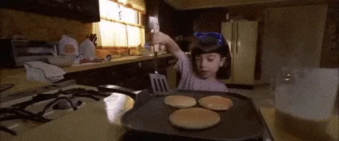 A split-screen image shows a young girl flipping pancakes on a griddle, then stacking them high on a plate. The top row…