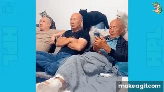 Three men sit on a couch, one in the middle holding a phone and looking at it, while the man on the right looks at him with…