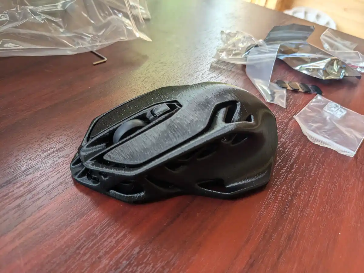 A black 3d-printed computer mouse sits on a wooden table, surrounded by plastic bags containing small parts and tools. The…