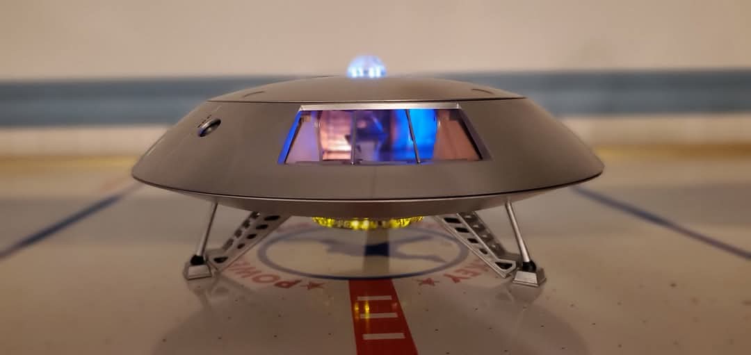 A small, silver, saucer-shaped model UFO with glowing blue lights on top and bottom rests on a landing pad marked with red…
