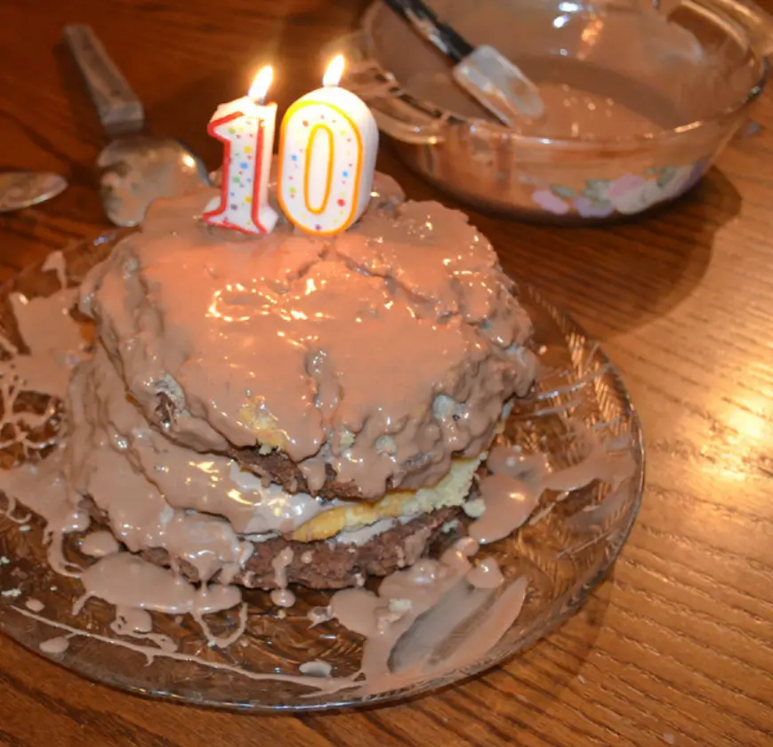 A homemade birthday cake with messy chocolate frosting, topped with two lit candles shaped like the number '10', sits on a…