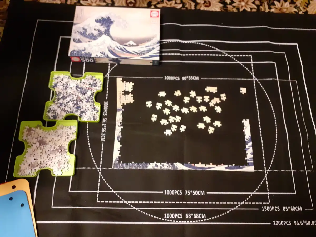 A jigsaw puzzle of 'The Great Wave off Kanagawa' is partially assembled on a black mat with white grid lines and text…
