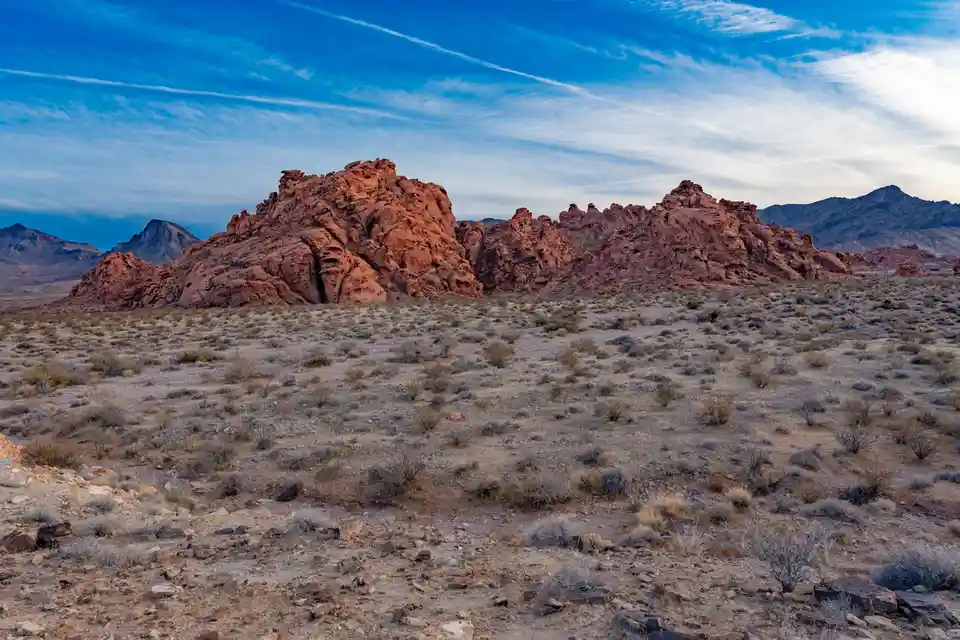 A wide desert landscape with red rock formations under a blue sky with wispy clouds. The foreground is sandy with sparse…