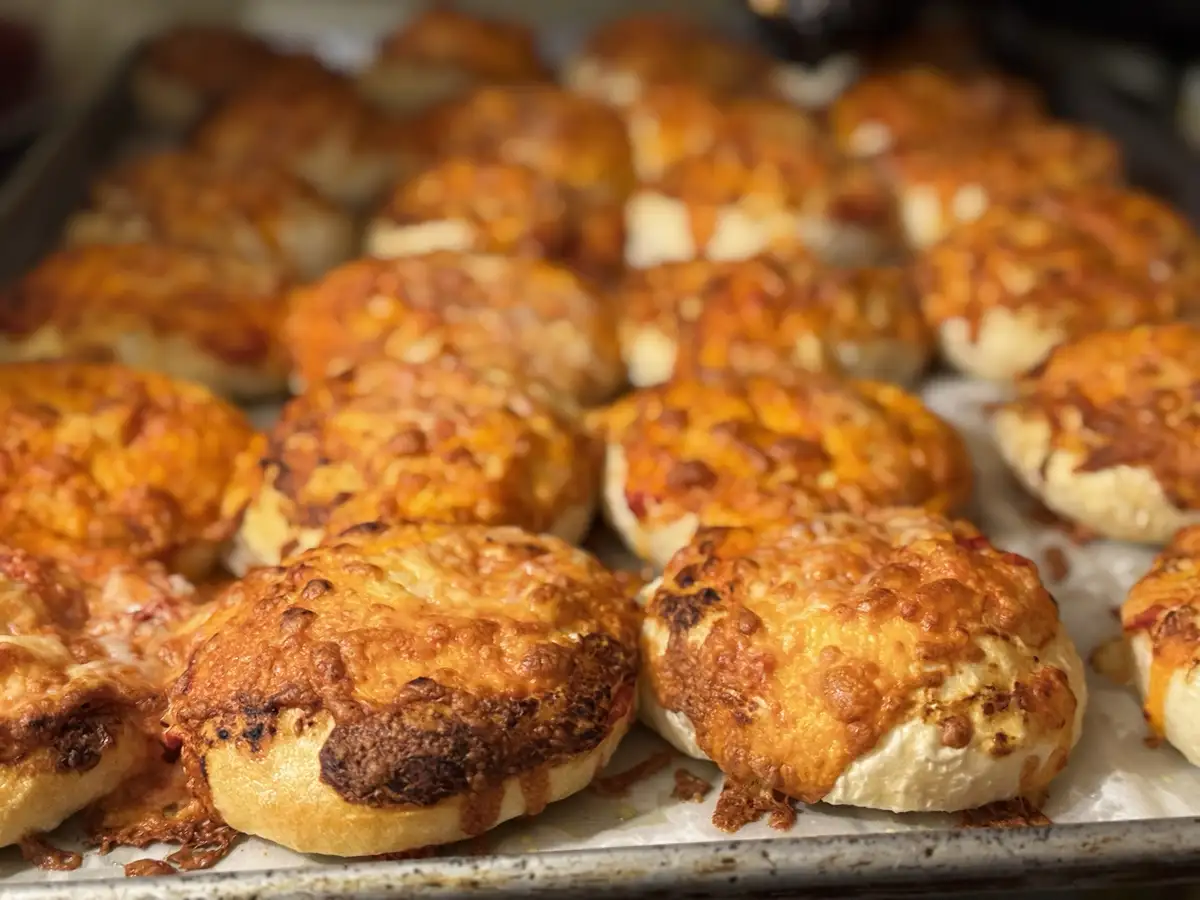 A close-up photo of a baking sheet filled with golden-brown, cheese-topped mini pizzas or bread rolls, some with slightly…