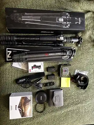 An overhead shot displays two camera tripods, their boxes, and various accessories like batteries, memory cards, lens caps…