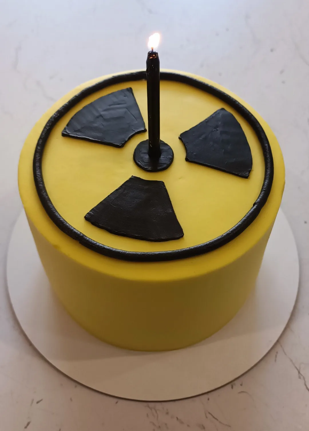 A round yellow cake decorated with a black radioactive symbol, featuring a single lit black candle placed in the center of…
