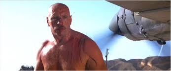 A shirtless, bald man with a mustache stares intensely at the camera while a large airplane engine looms behind him…