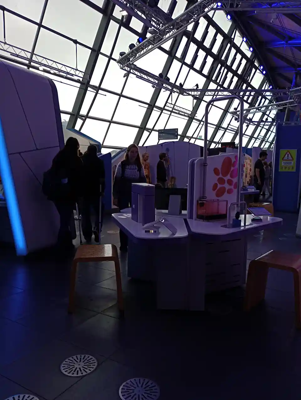 A photo of a science museum exhibit with purple tables, stools, and interactive displays under a glass roof. Several people…
