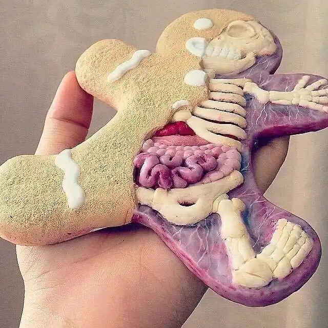 A hand holds a gingerbread man cookie that has been hollowed out and filled with detailed edible anatomical models…