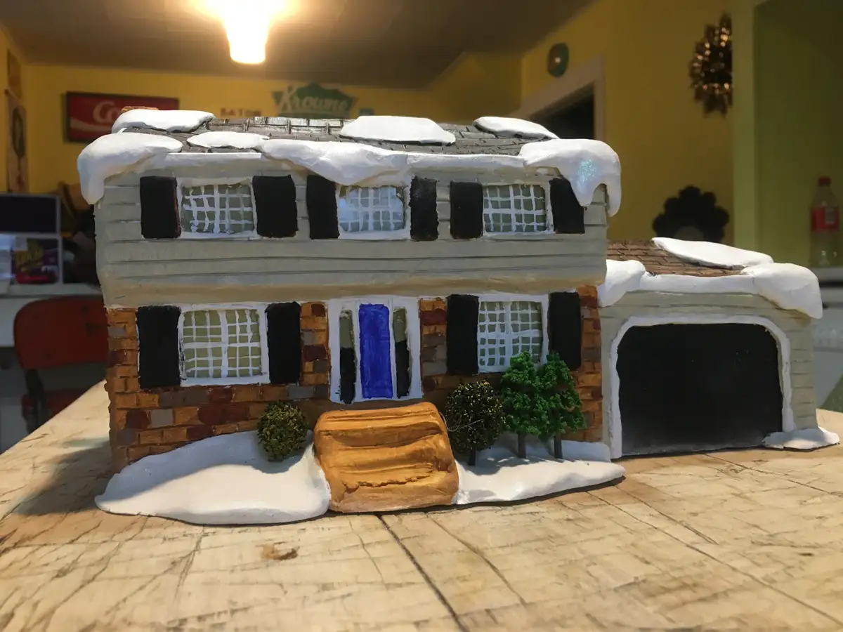 A handmade gingerbread house model sits on a wooden table, decorated with white icing to look like snow on the roof and…