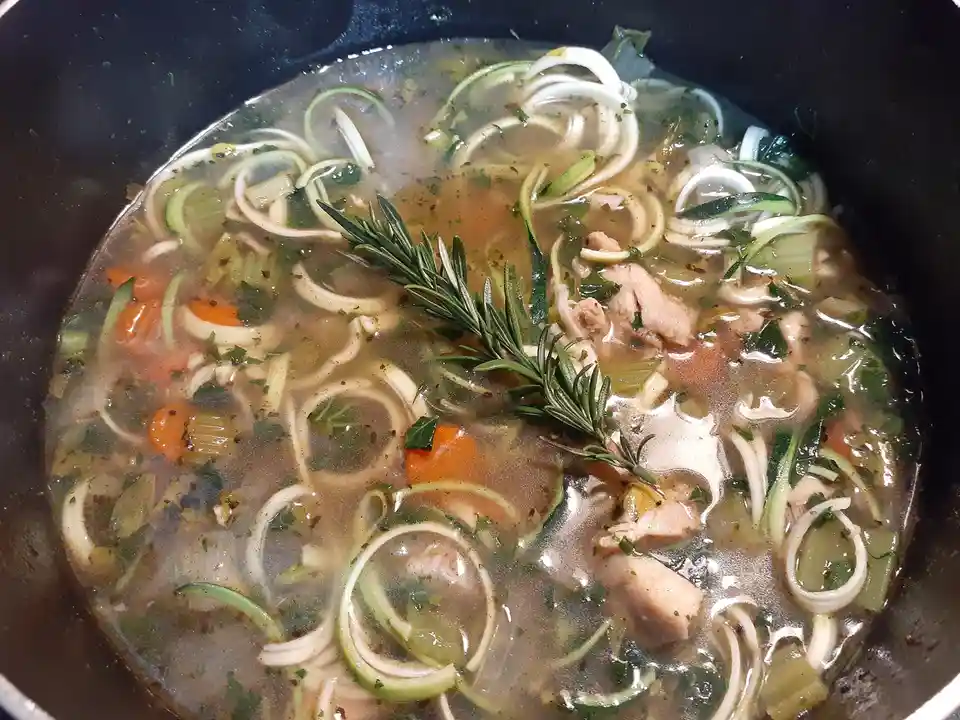 A close-up photo of a pot filled with clear broth, chunks of chicken, sliced carrots, and spiralized zucchini noodles…