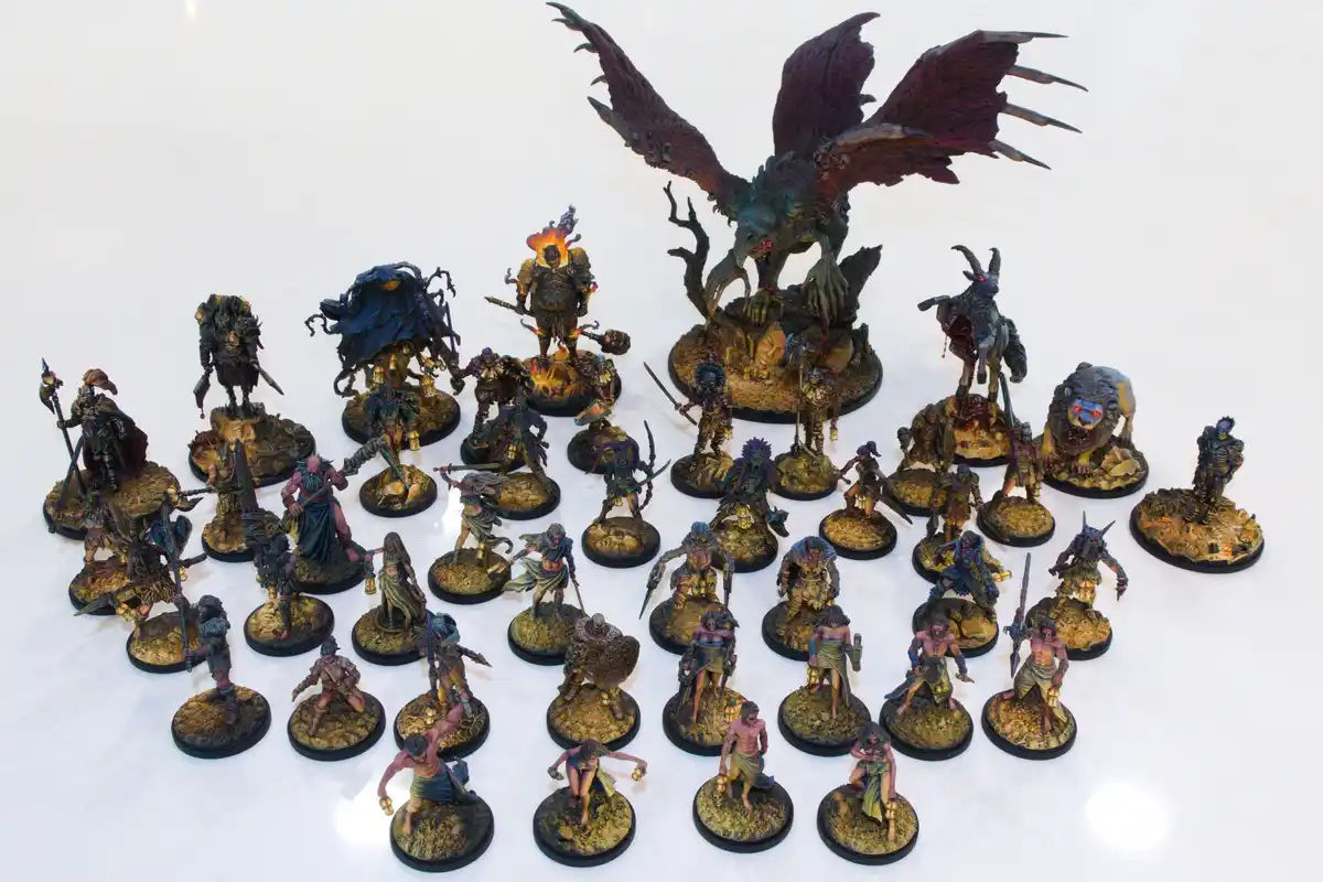 An overhead shot displays a large collection of intricately painted fantasy miniatures on circular bases, arranged on a…
