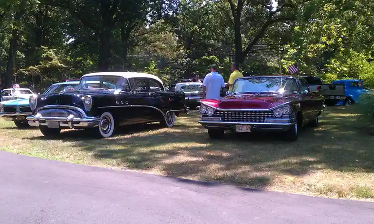 A photo of several classic American cars parked on grass under trees, with two men standing near a red 1959 Cadillac with…