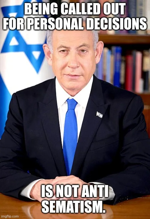 A man in a dark suit and blue tie sits at a desk with his hands clasped, looking serious. Behind him is an Israeli flag and…