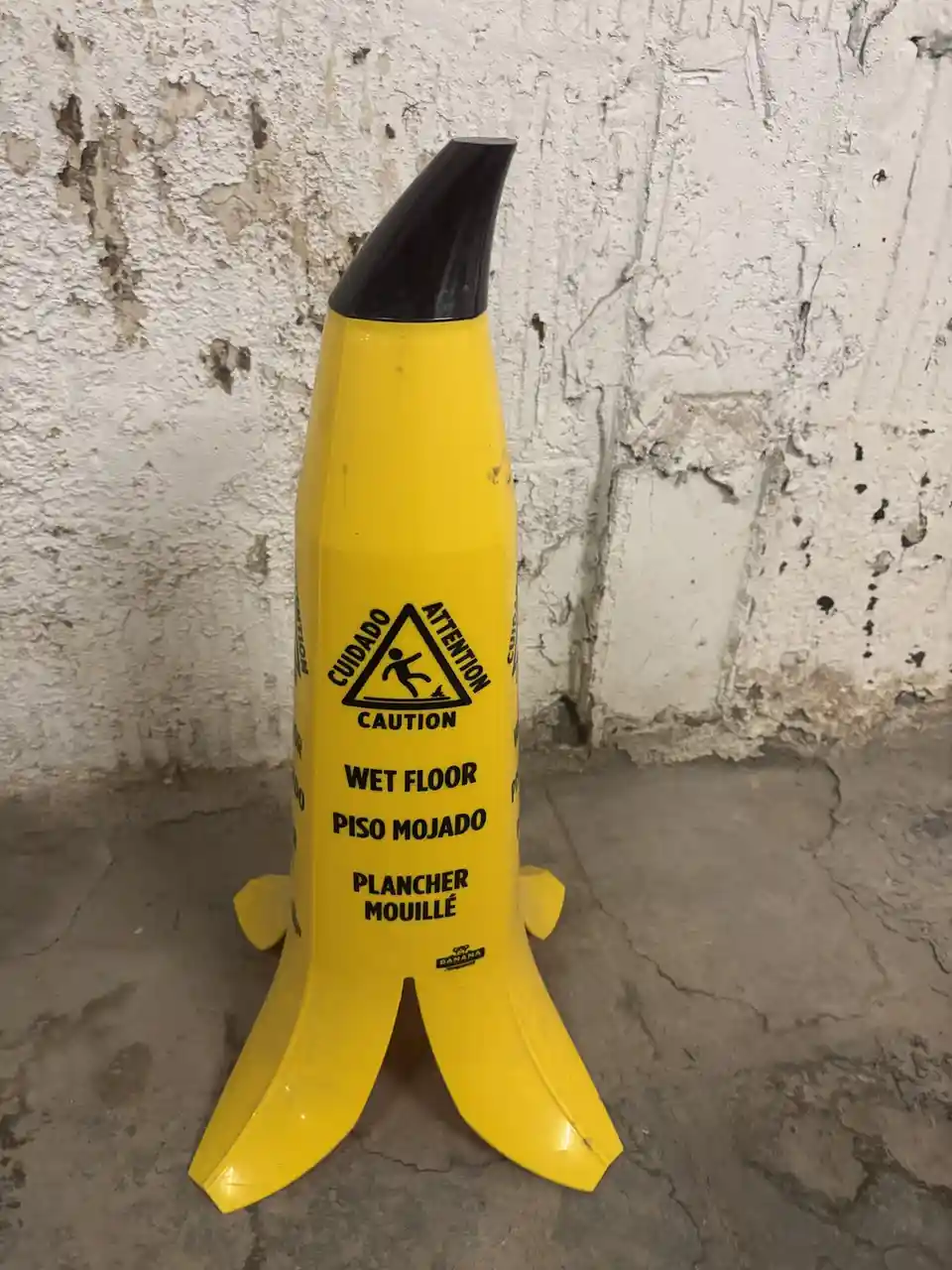 A yellow, banana-shaped caution sign stands on a cracked concrete floor against a weathered white wall. The sign features a…