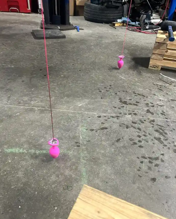 Two pink, bulbous sex toys hang by thin pink strings from above in a cluttered garage with concrete floor, tools, and a tire…