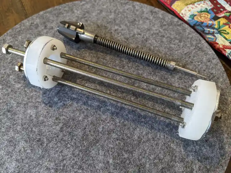 A 3D-printed mechanical device with three threaded rods extending from a white circular base, resting on a gray felt…