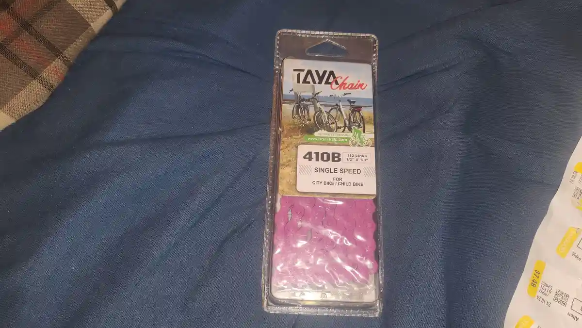 A package of a bright pink TAYA bicycle chain, labeled '410B SINGLE SPEED FOR CITY BIKE / CHILD BIKE', sits on a blue fabric…