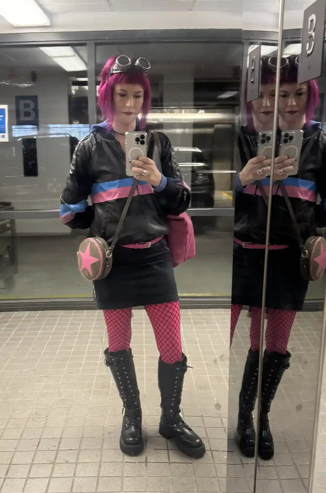 A person with bright pink hair and goggles takes a mirror selfie in a parking garage, wearing a black jacket with pink and…