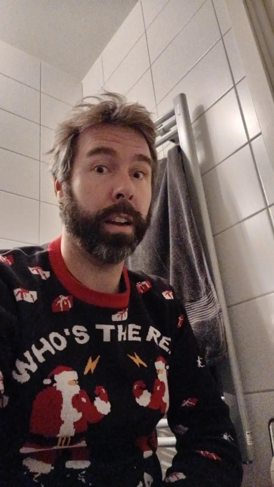 A man with a beard and messy hair takes a selfie in a bathroom wearing a black Christmas sweater with red trim and cartoon…