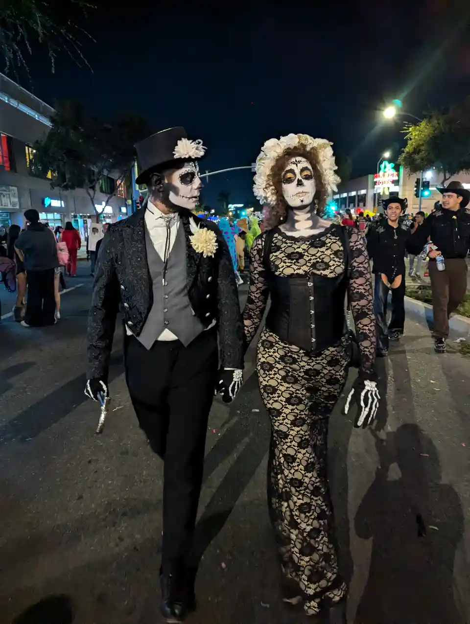 Two people dressed as sugar skulls, wearing elaborate black lace costumes and skull makeup, walk hand-in-hand down a city…