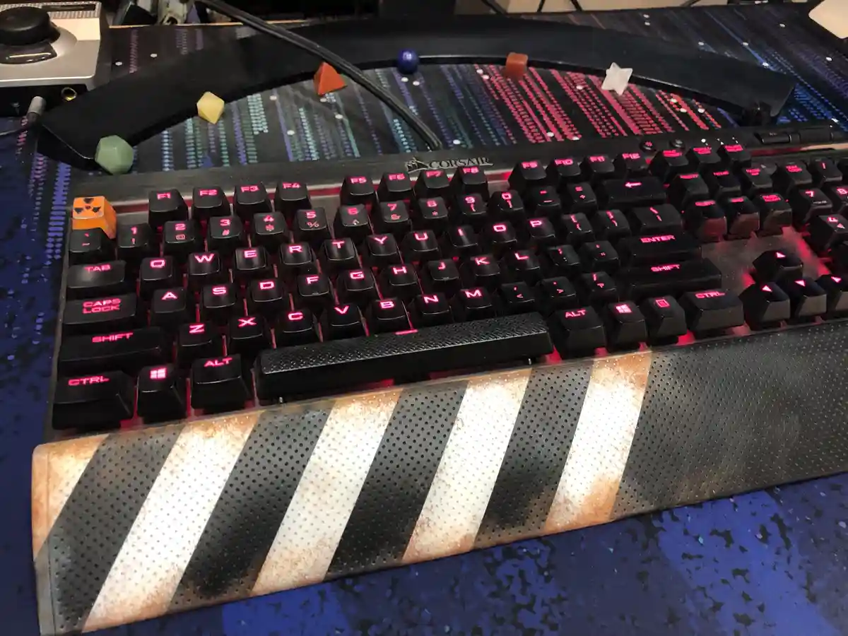 A black Corsair mechanical keyboard with red backlighting sits on a blue patterned desk mat. The keyboard's wrist rest has…