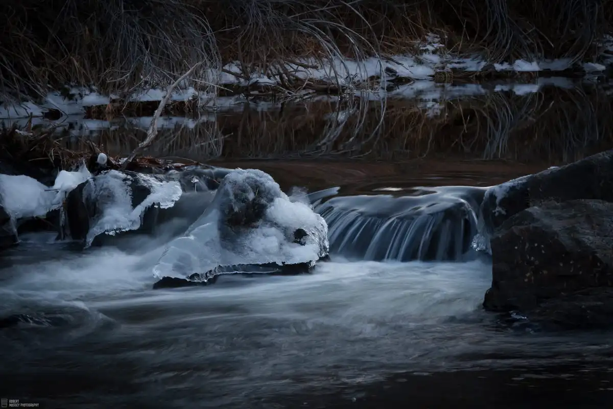 A long-exposure photograph of a dark, icy stream with water flowing over rocks covered in ice and snow, surrounded by bare…