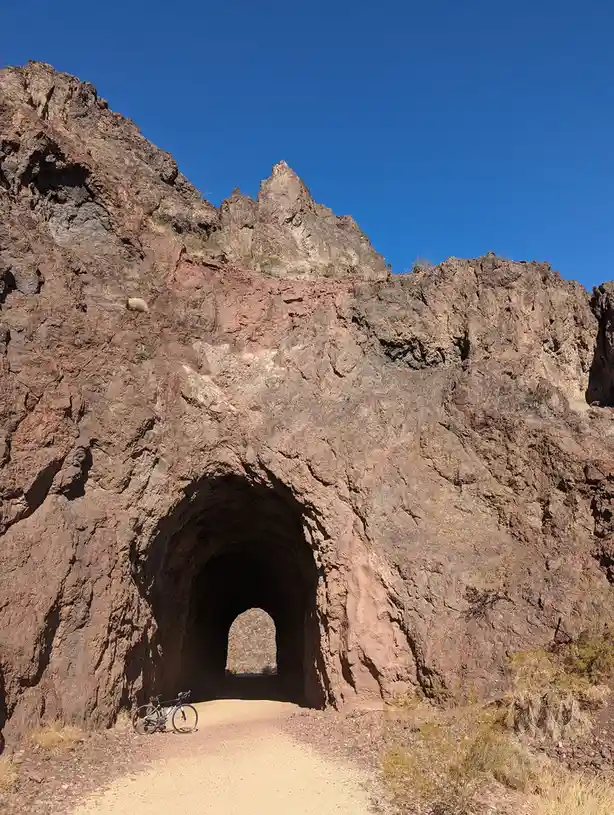 A black road bicycle is parked on a dirt path at the entrance of a natural rock tunnel carved into a reddish-brown cliff…