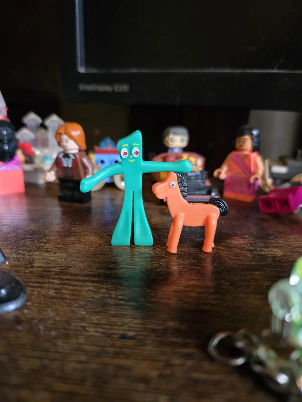 A teal, stick-figure-like toy with red eyes and a yellow smile stands next to an orange plastic horse on a wooden desk. The…