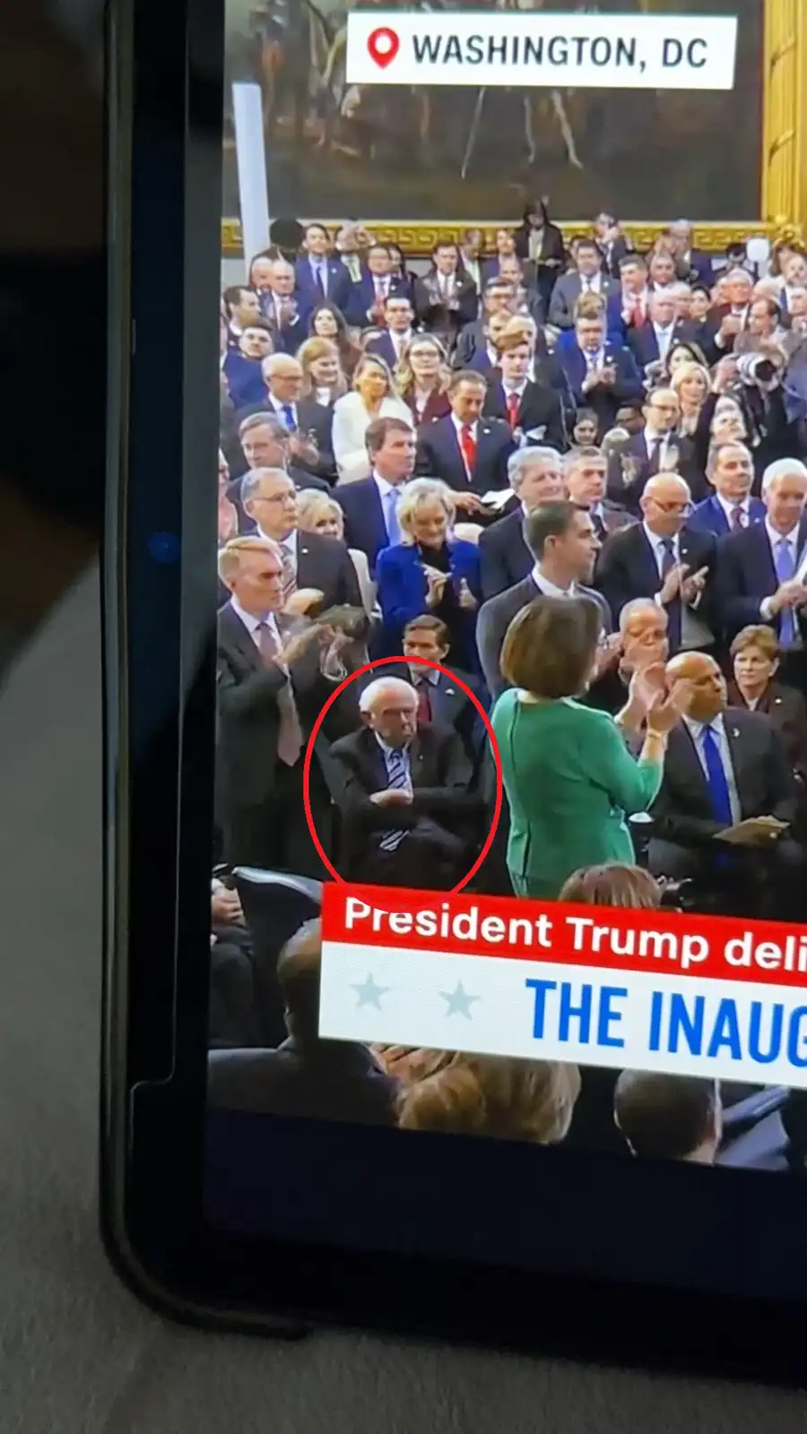 A smartphone screen displays a televised inauguration event in Washington, DC, showing a crowd of formally dressed…