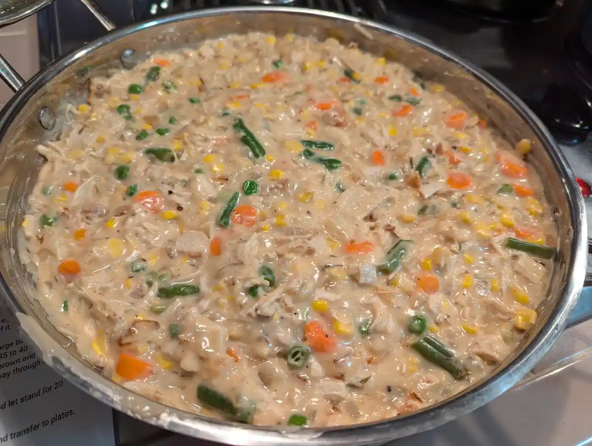 A close-up photo shows a large stainless steel skillet filled with a creamy, thick stew containing shredded chicken, corn…