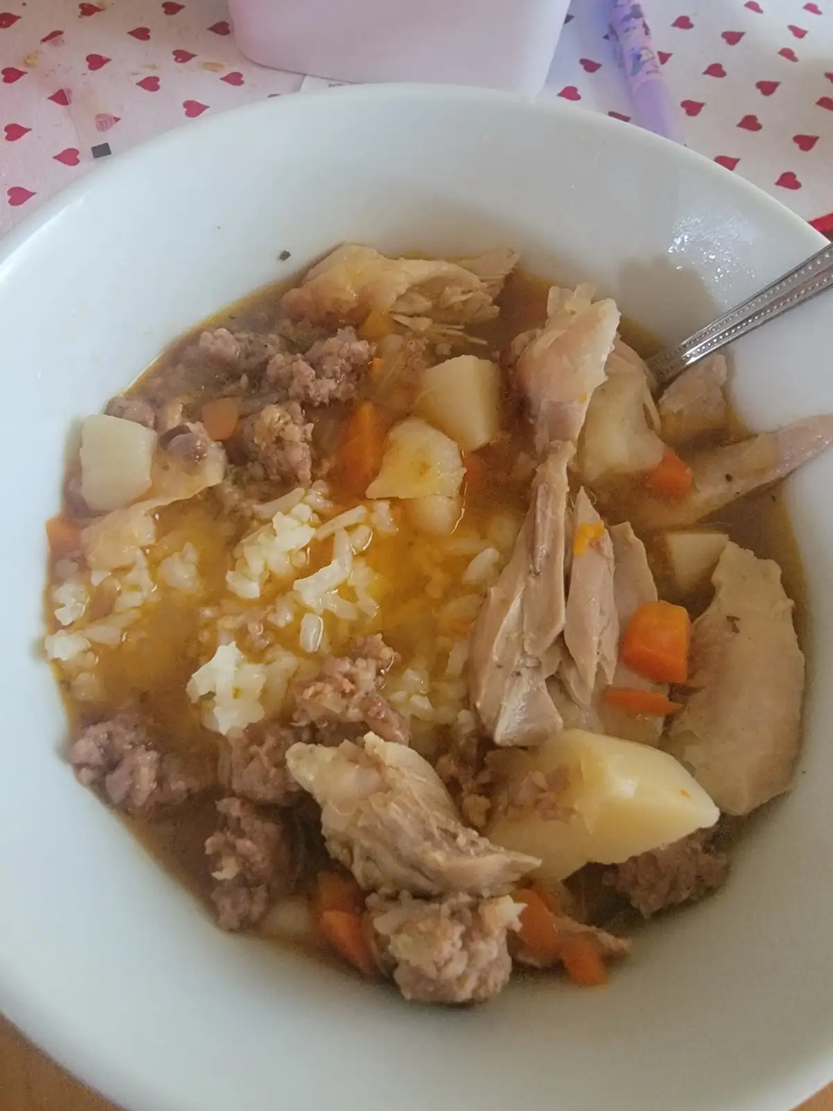 A white bowl filled with a stew-like dish containing shredded chicken, ground meat, diced potatoes, and carrots in a brown…