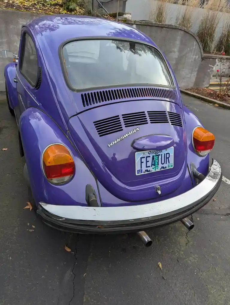 A purple Volkswagen Beetle is parked outdoors, showing its rear with a custom Oregon license plate that reads 'FEATUR'. The…
