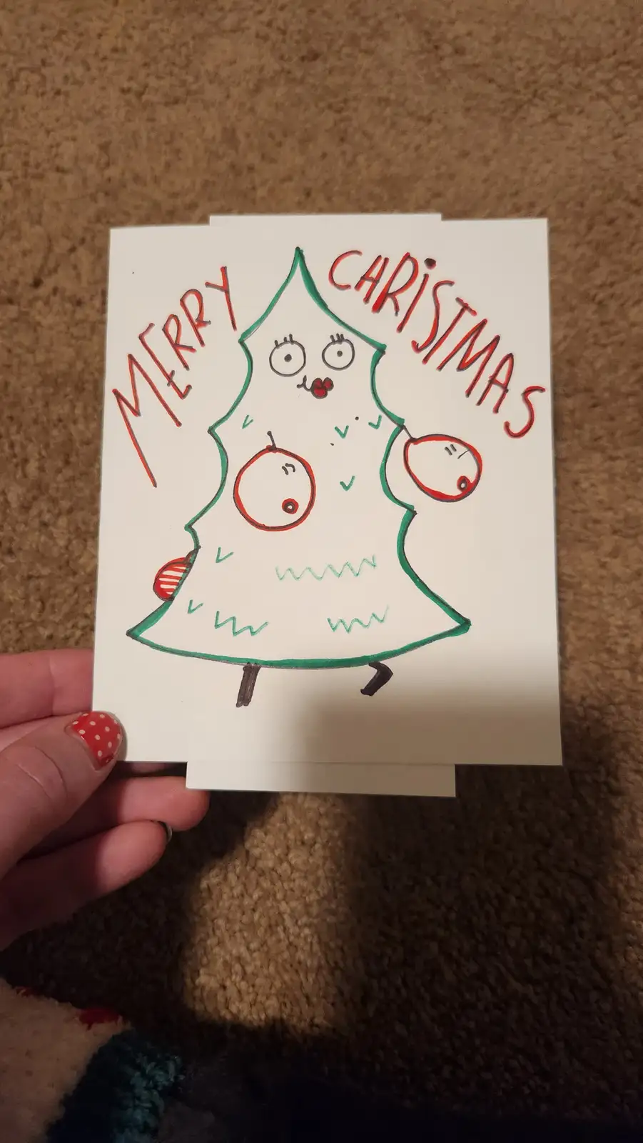 A hand holds a handmade Christmas card featuring a cartoonish, anthropomorphic Christmas tree with a surprised or shocked…