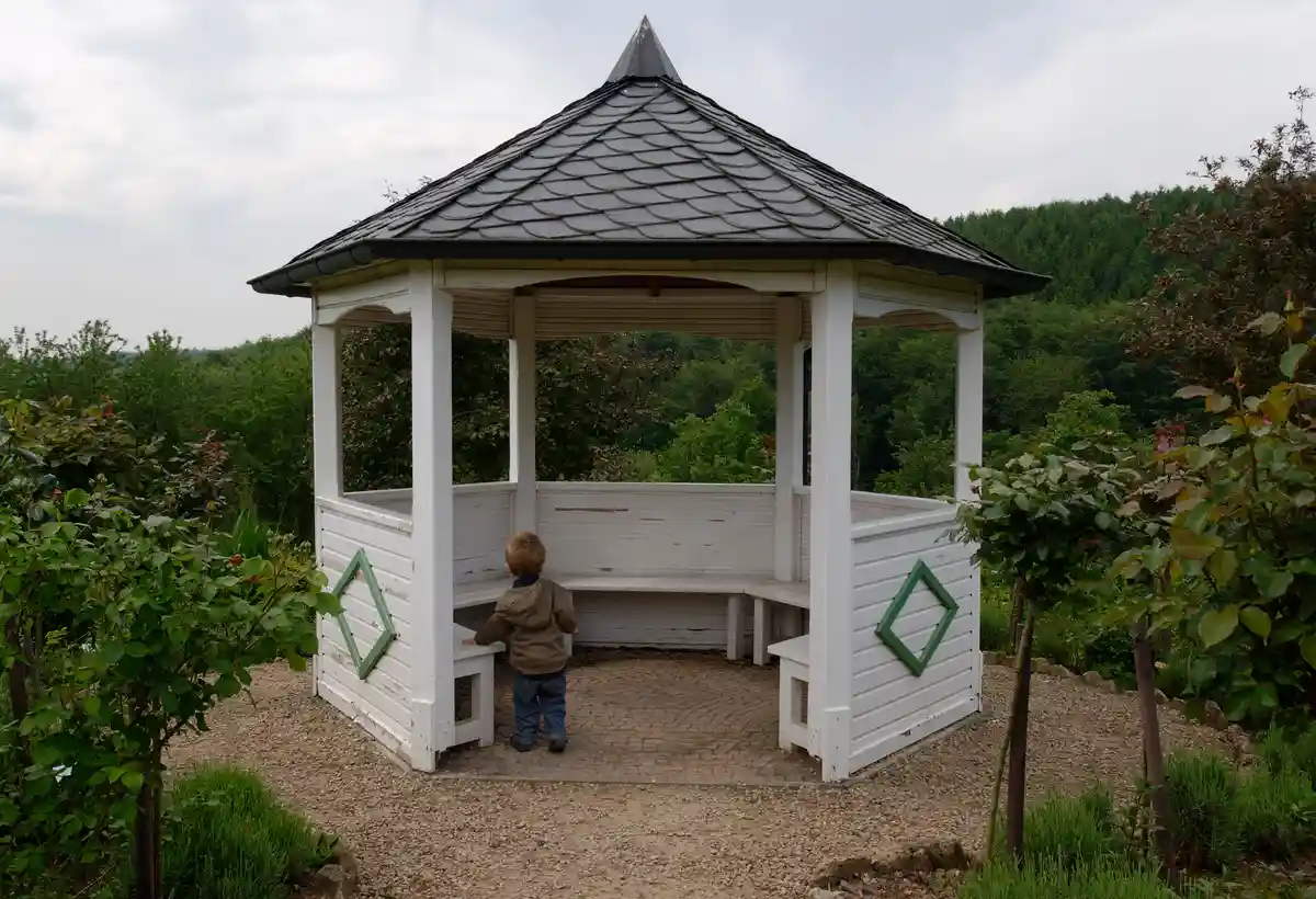 A young child in a brown jacket and jeans stands inside a white wooden gazebo with a dark shingled roof, looking out at a…