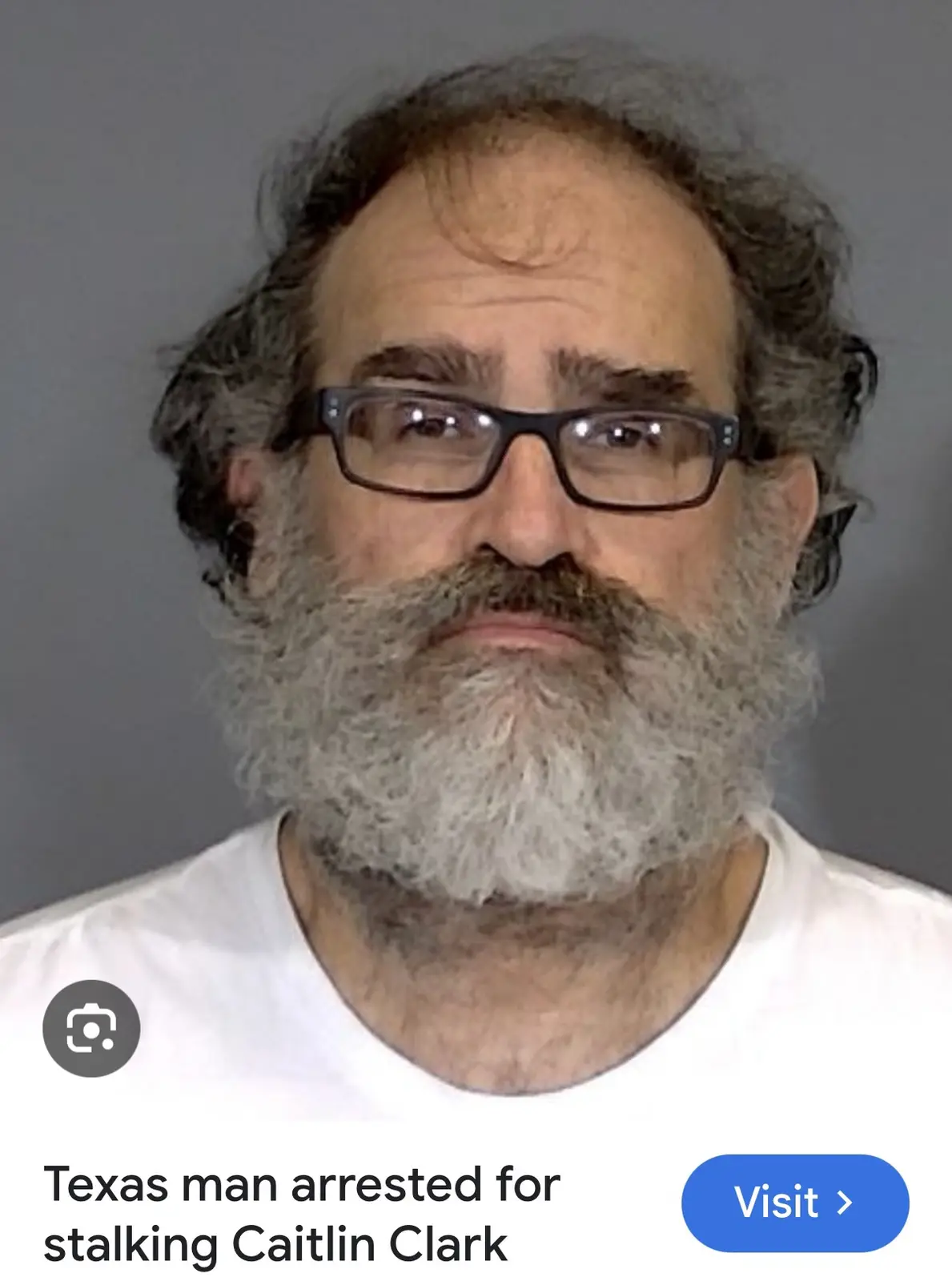 A mugshot of a middle-aged man with a full gray beard, glasses, and a white t-shirt against a gray background. The image has…