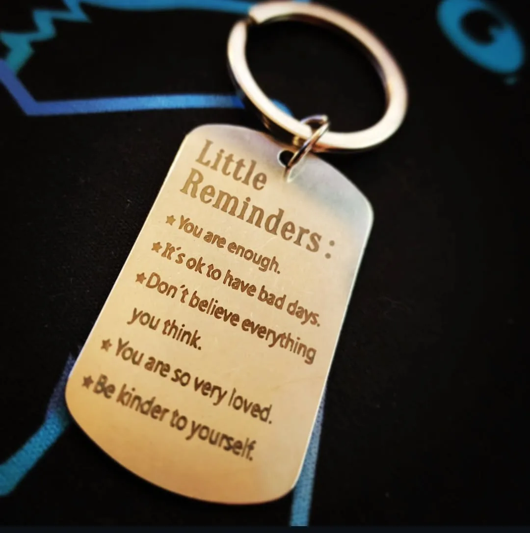 A close-up photo of a brushed metal dog tag keychain with engraved motivational phrases under the heading 'Little…