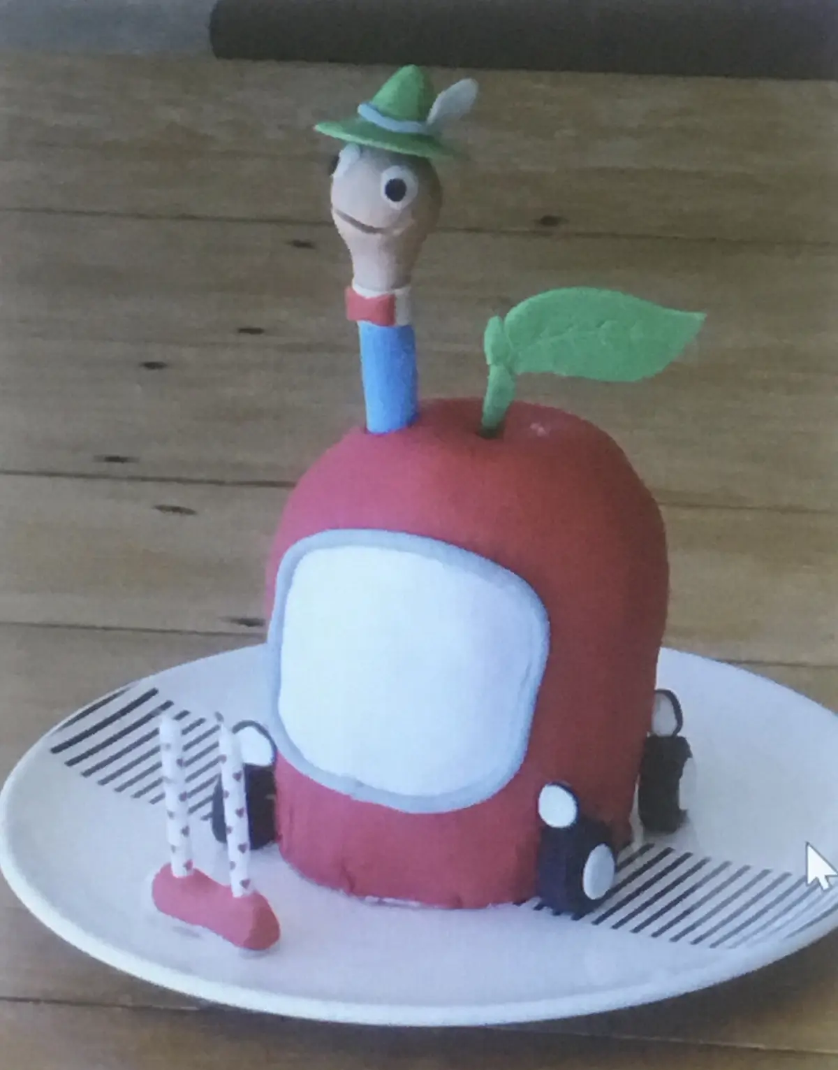 A cake shaped like a red apple with a green leaf and a cartoon character with a green hat sticking out of the top, sitting…