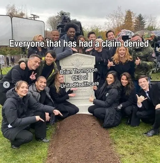 A group of smiling people posing around a freshly dug grave with a tombstone. The tombstone has text identifying Brian…