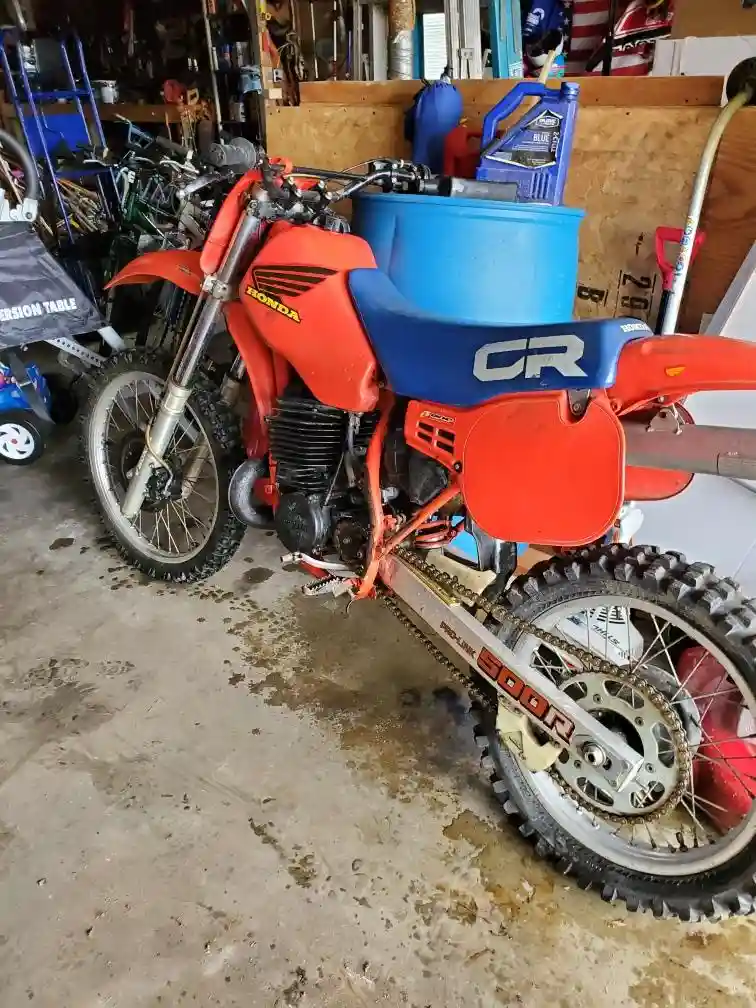 A red and blue Honda CR dirt bike is parked in a cluttered garage, with a large blue bucket strapped to its seat. The bike…