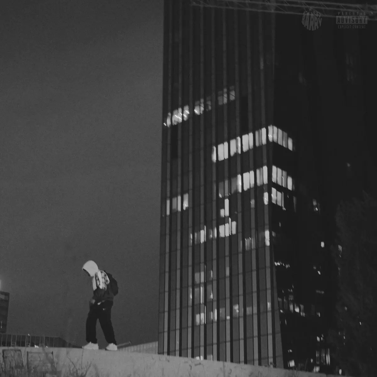 A black and white photo shows a person in a hoodie walking away from the camera on a rooftop at night, with a tall, lit…