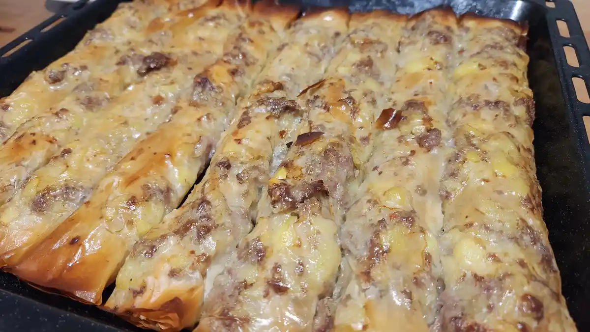 A close-up photo of a black baking tray filled with rows of golden-brown, cheesy, meat-filled rolled phyllo dough…